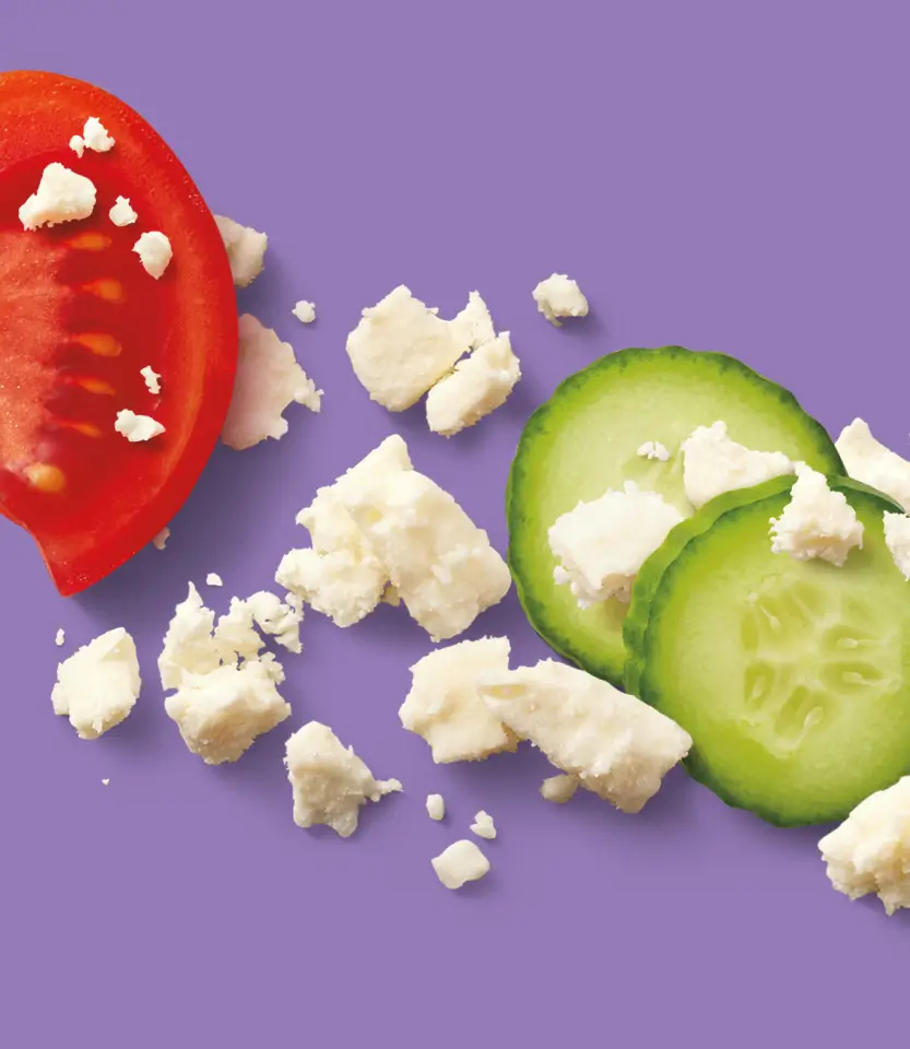 Sliced red and green tomatoes with crumbled white cheese on purple background, arranged in flat lay style.