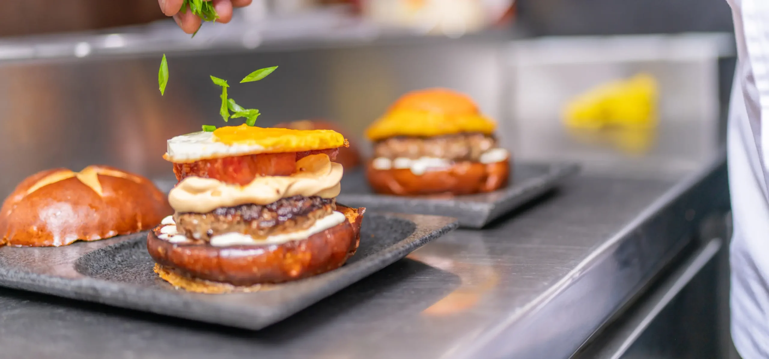 Gourmet burgers on pretzel buns with patties, cheese, and orange garnish being plated on slate, with fresh herb garnish being added by hand.