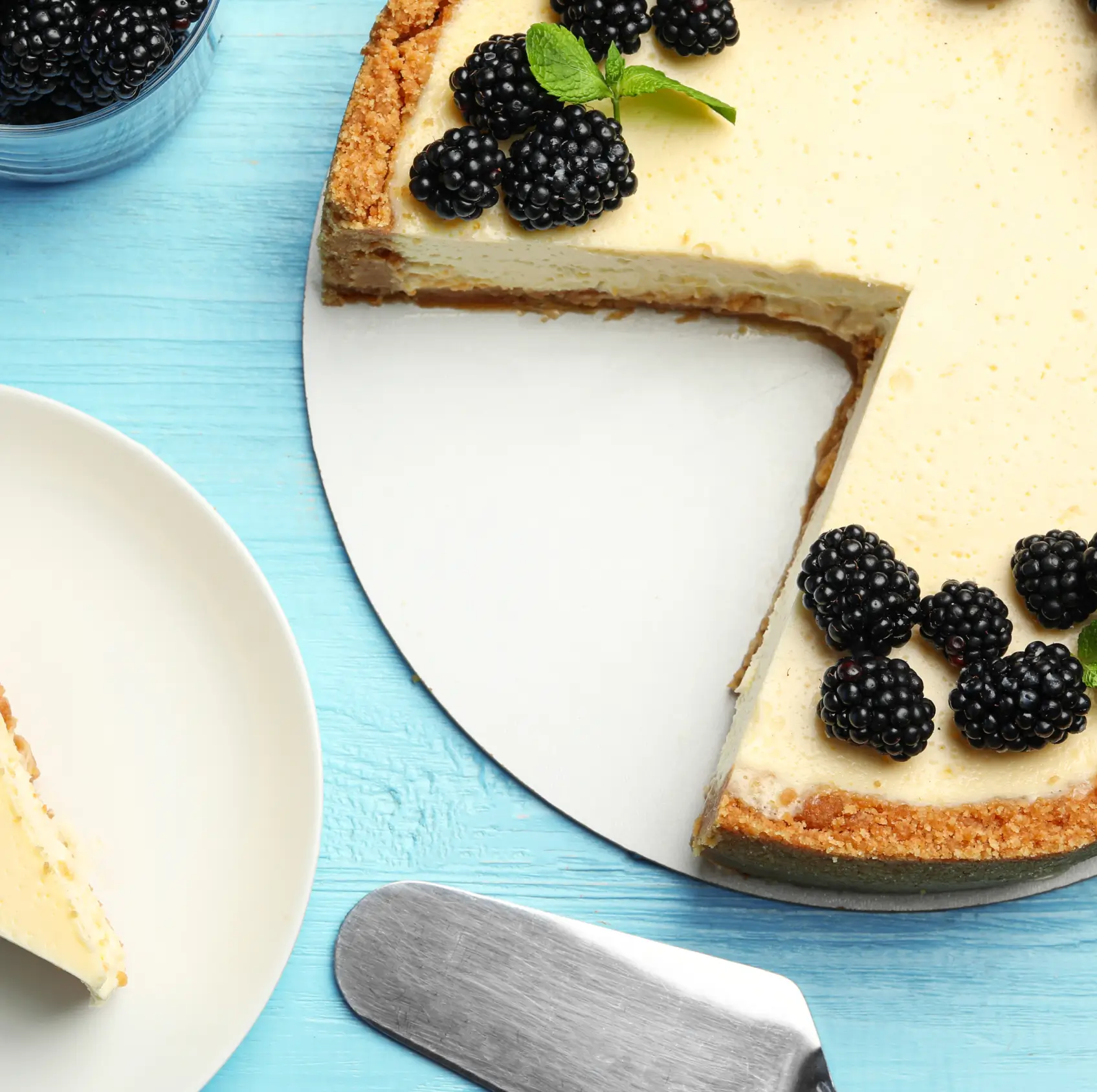 Cheesecake with graham cracker crust decorated with fresh blackberries and mint, displayed on white plate with blue wooden surface and cake server.