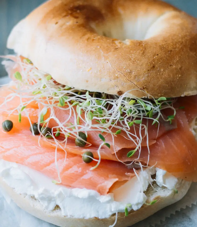 Toasted bagel sandwich with smoked salmon, cream cheese, sprouts, and capers on a light blue surface.