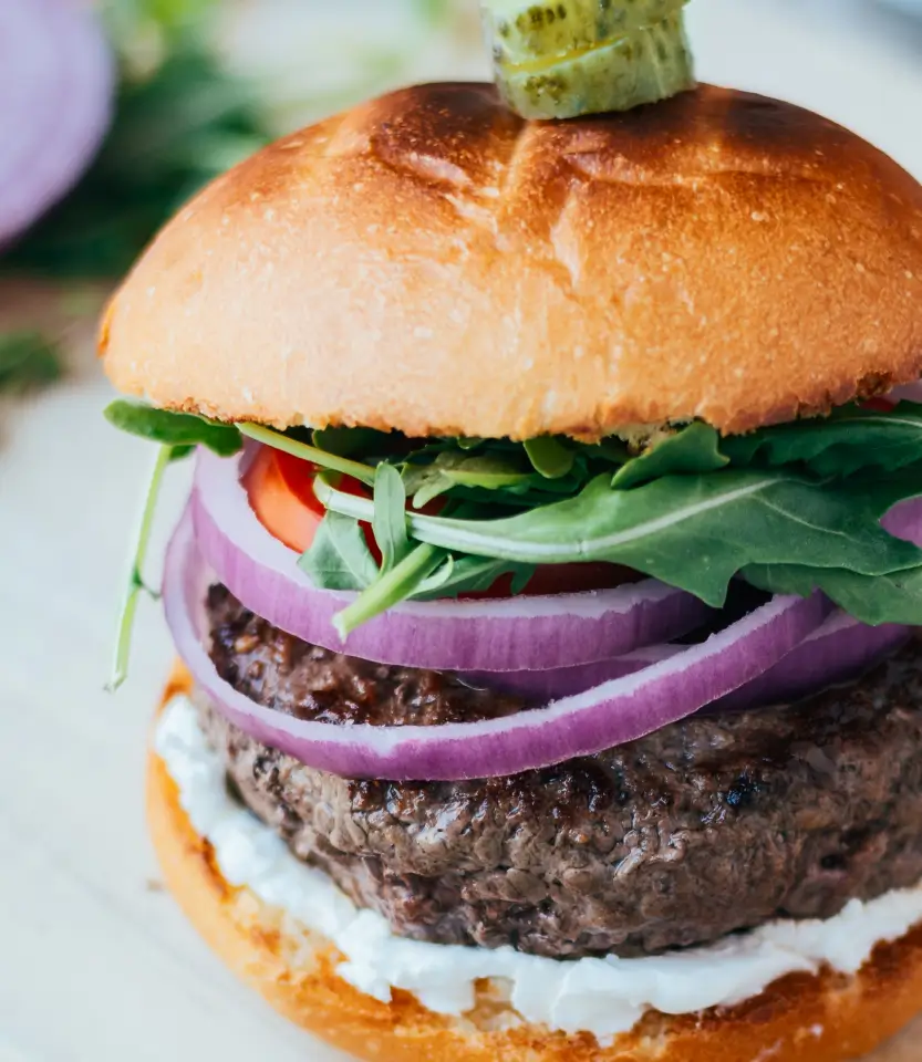 Gourmet burger with toasted bun, beef patty, white sauce, purple onion, green arugula, tomato, and pickle garnish on top.