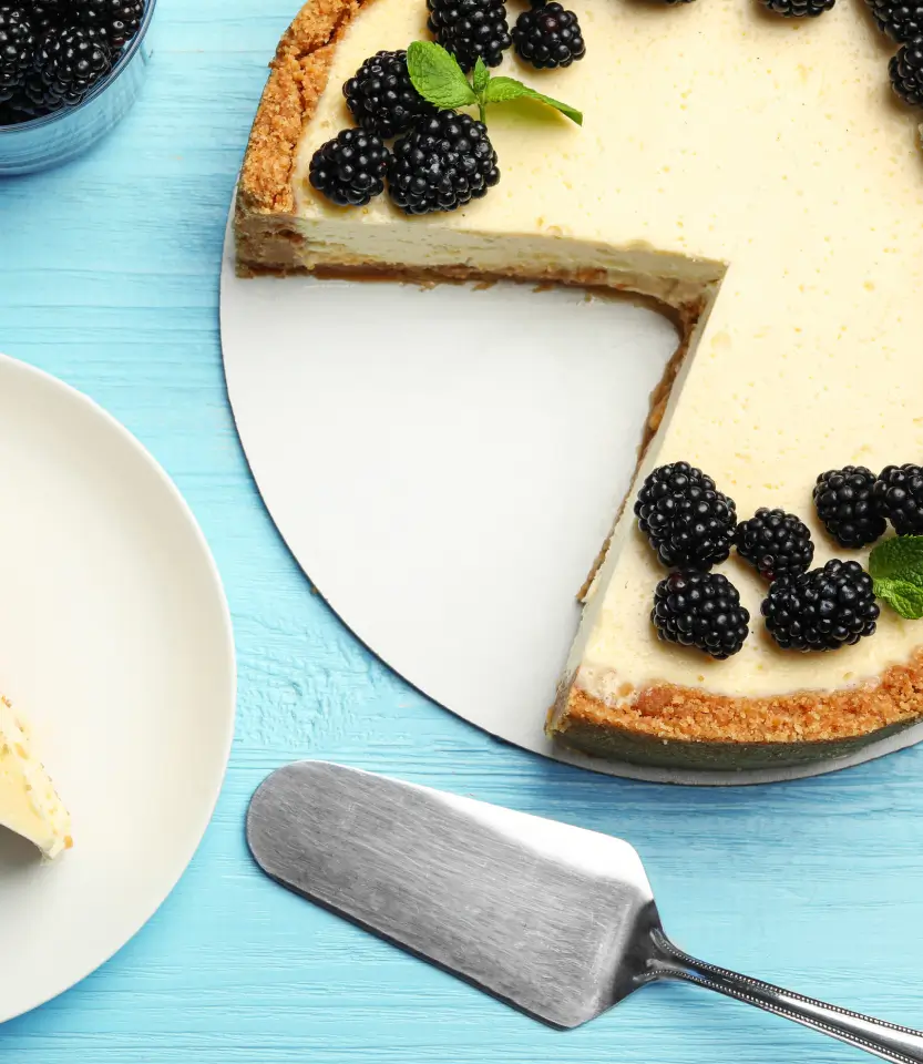 Blackberry cheesecake with graham cracker crust on blue wooden surface, one slice removed, served with cake server and empty plate.