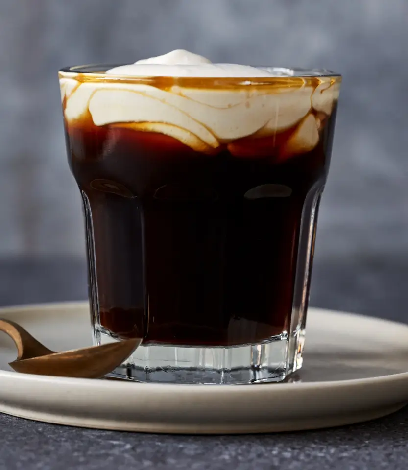 Tall glass of dark cola with ice and whipped cream on top, served on a white plate with a stirrer.