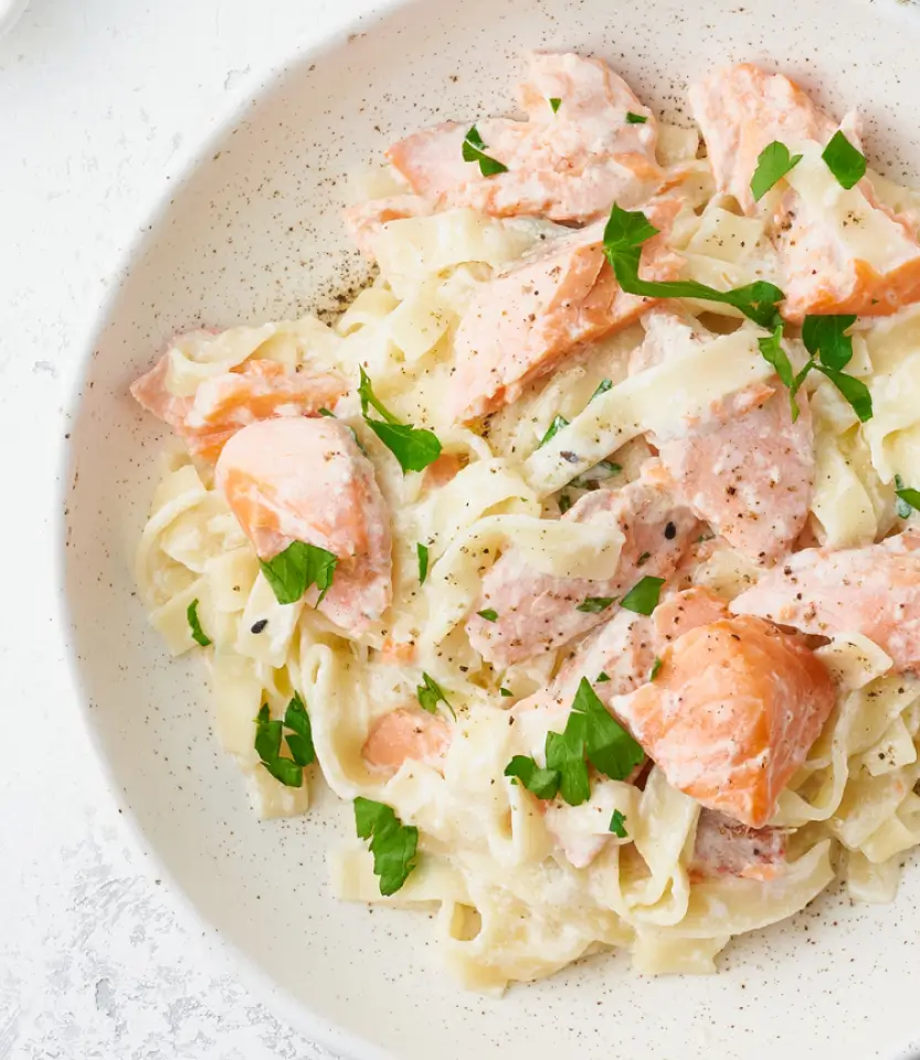 Creamy pasta dish with smoked salmon, wide ribbon noodles, black pepper, and fresh parsley garnish on a speckled white plate.