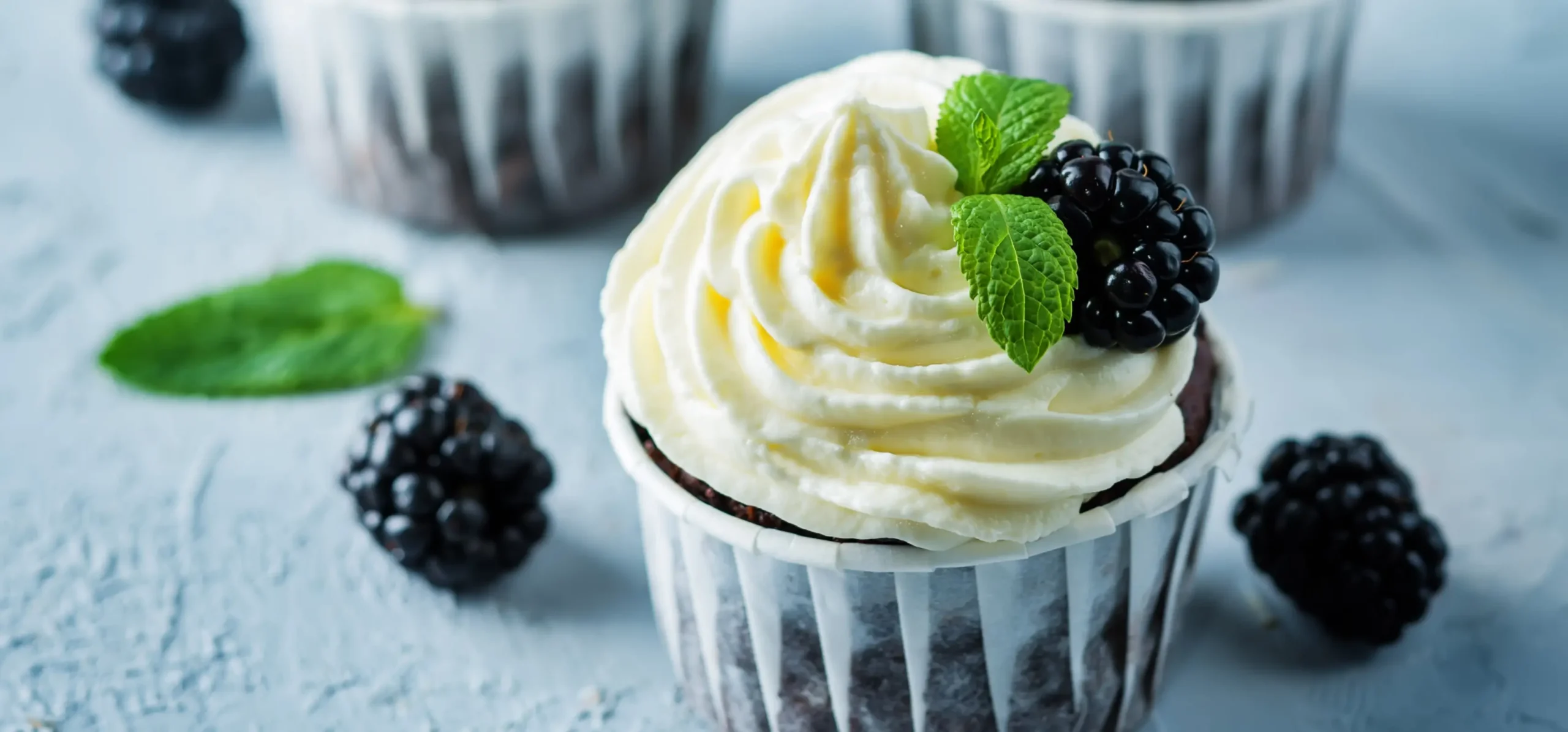 Chocolate cupcake with cream frosting, topped with fresh blackberry and mint leaf, surrounded by scattered blackberries on a light blue surface.