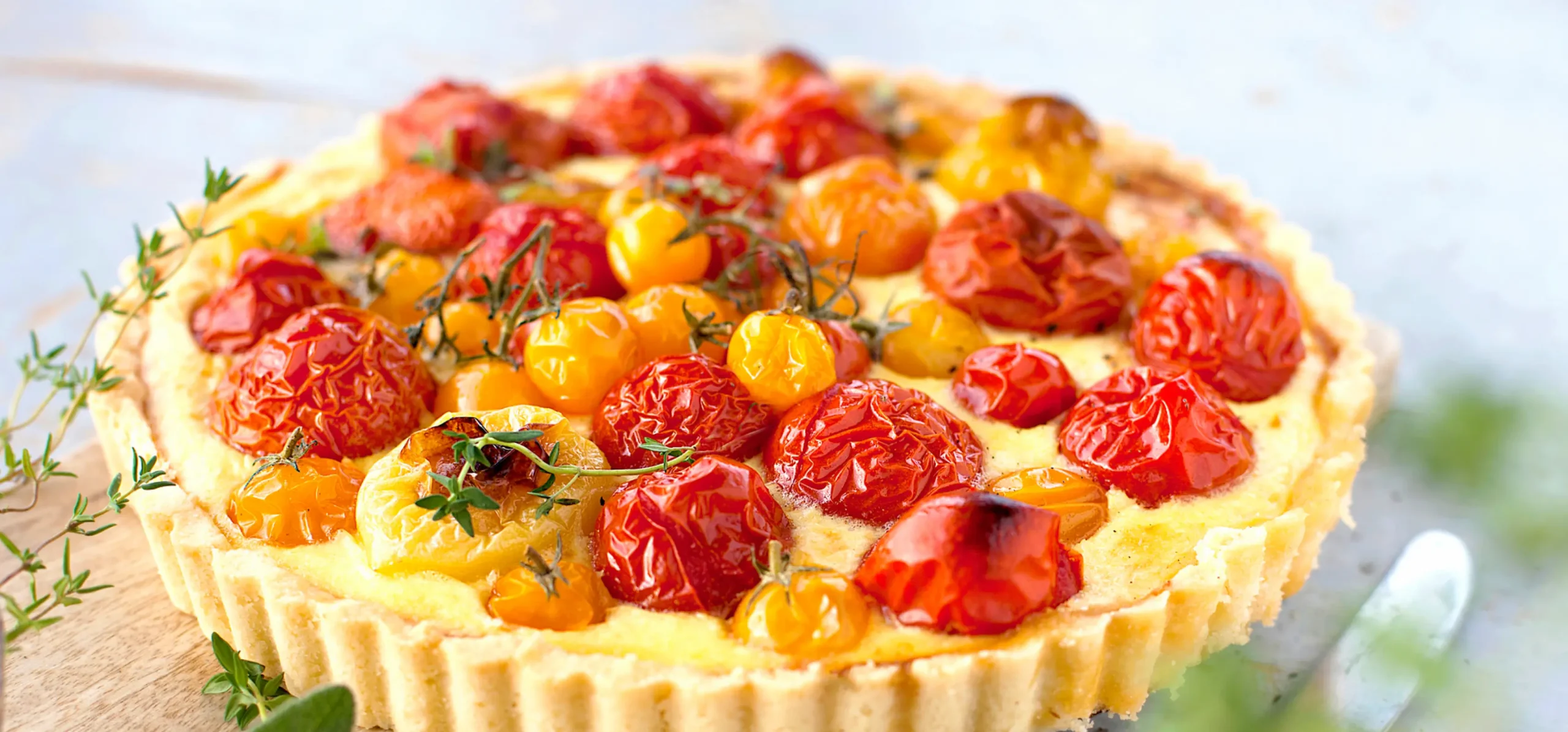Savory tart with roasted red and yellow cherry tomatoes on creamy filling in pastry crust, garnished with fresh thyme sprigs.