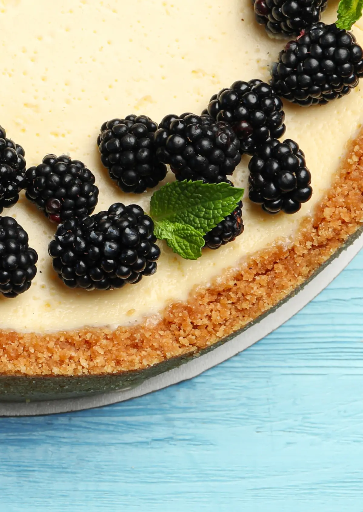 Blackberry cheesecake with graham cracker crust on blue wooden surface, topped with fresh blackberries and mint leaf.
