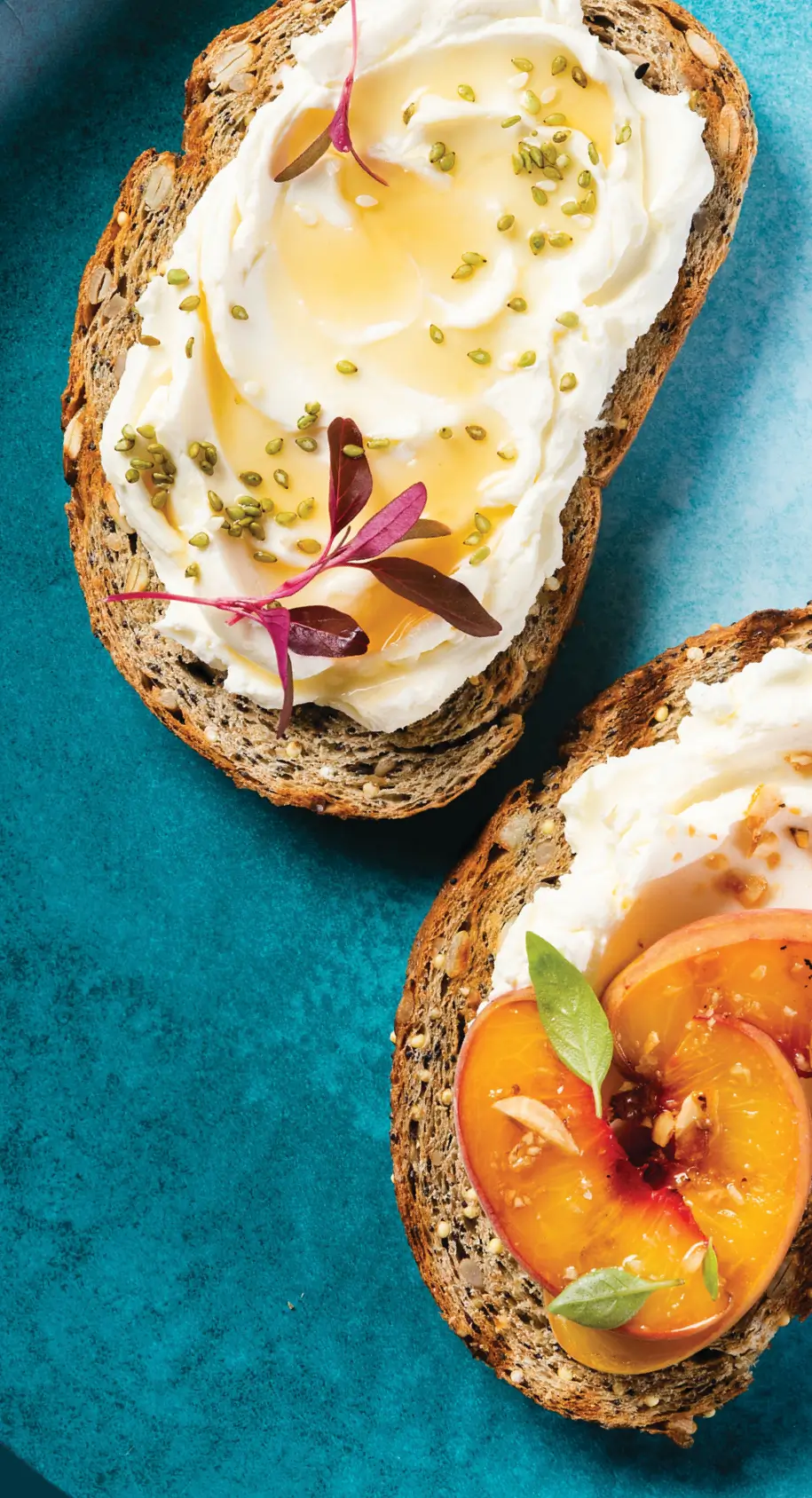 Two open-faced sandwiches on seeded bread with cream cheese, topped with pumpkin slices, pistachios, and edible flowers on a teal background.